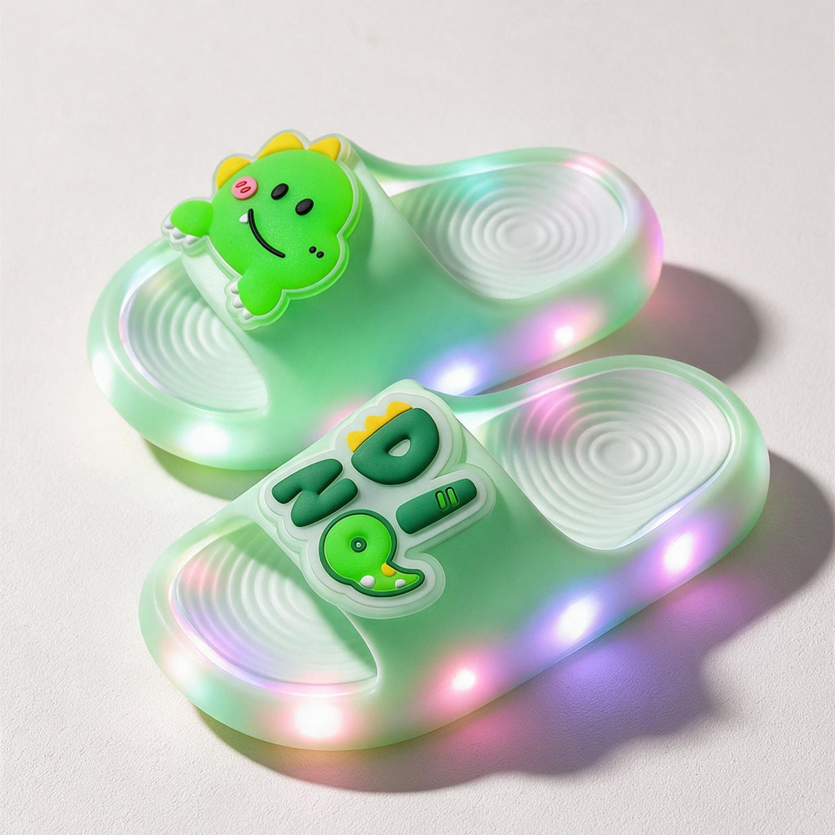 A pair of green Dino STEPGLO slides, one strap with a smiling dinosaur and the other with DINO in raised letters, LEDs lit around both soles.