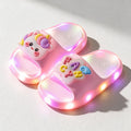 A pair of pink STEPGLO light-up slides in sunlight, one strap with a unicorn face and the other with FANCY in raised letters, LEDs lit around both soles.