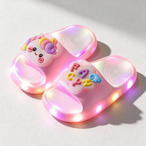A pair of pink STEPGLO light-up slides in sunlight, one strap with a unicorn face and the other with FANCY in raised letters, LEDs lit around both soles.